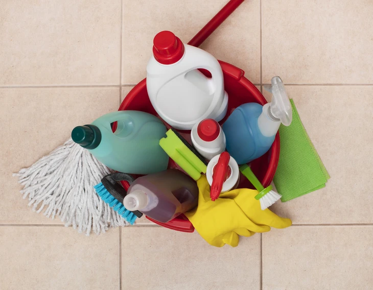 Scène intérieure sur carrelage beige montrant un seau rouge contenant divers produits et outils de nettoyage ménager