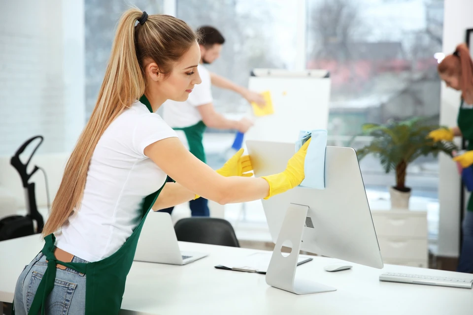 Femme blonde en uniforme de nettoyage blanc et tablier vert pulvérise un produit désinfectant sur écran d'ordinateur iMac dans bureau moderne