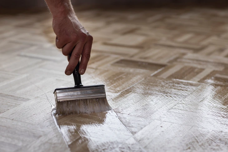 Une main applique un produit de finition transparent sur un parquet en bois naturel à l'aide d'un pinceau plat professionnel