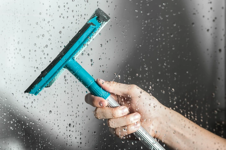 Une main tient une raclette de nettoyage turquoise sur une surface vitrée couverte de gouttelettes d'eau