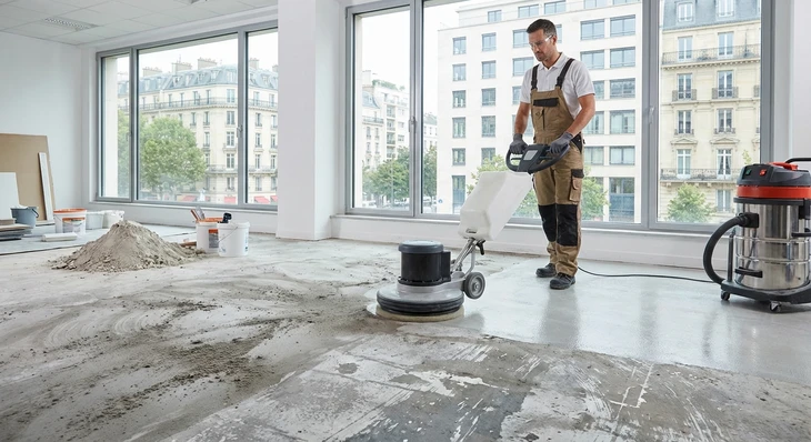 Photographie en intérieur d'un chantier en cours de finition montrant un professionnel du nettoyage en tenue de travail utilisant une machine de décapage ou ponçage des sols pour enlever les résidus de chantier, marques et salissures tenaces sur une surface de sol partiellement traitée, avec des zones encore visiblement souillées avant nettoyage.