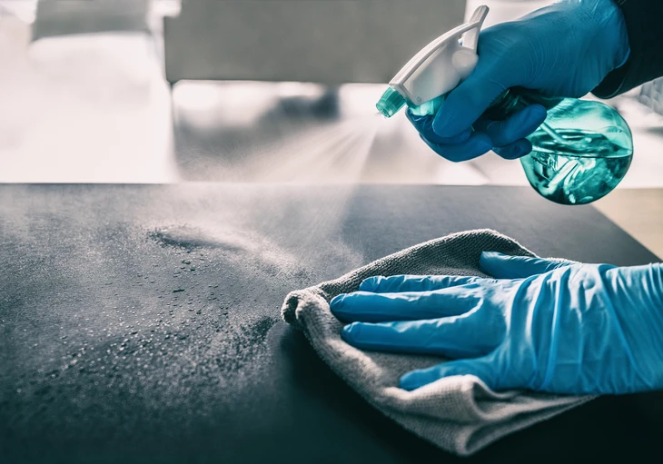 Personne portant des gants de protection nitrile bleus effectuant un nettoyage et désinfection de surface avec pulvérisateur et chiffon microfibre