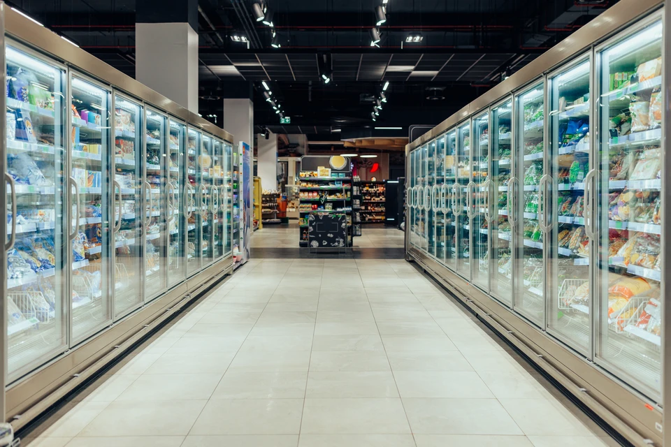 Allée de supermarché vue depuis un caddie, rayonnages hauts chargés de produits alimentaires emballés des deux côtés, sol carrelé blanc, éclairage néon au plafond, perspective centrale menant vers le fond du magasin